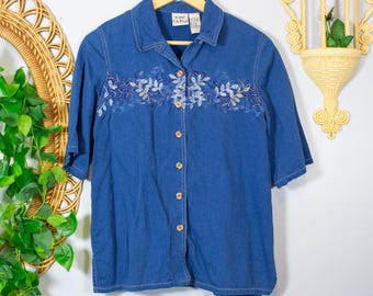 May include: A blue short-sleeved button-down shirt featuring a collar and a horizontal band of white and light blue leaf embroidery across the chest. The shirt has a front button closure and a label reading Koret City Blues.