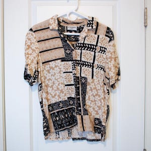 May include: A short-sleeved button-down shirt with a beige, black, and white floral and geometric pattern. The shirt is hanging on a white hanger against a white door. The shirt has a collared neckline and a button-up front.
