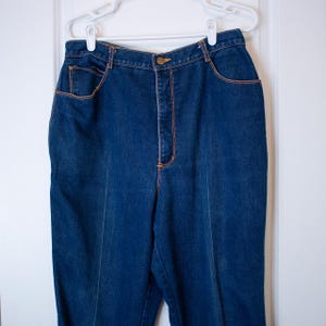 May include: Dark blue denim pants with orange stitching and a button closure. The pants have front and back pockets and are hanging on a white hanger. The jeans are a classic style.