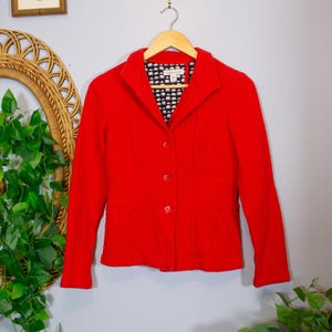 May include: A bright red textured blazer with a notched collar and three front buttons. The interior lining features a repeating black and white elephant pattern. The garment is displayed on a wooden hanger against a light gray wall background.