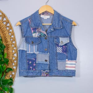 May include: A sleeveless denim vest with patchwork details. The vest features various fabric patches with floral, striped, and solid patterns. It has a button-up front and two chest pockets. The vest is hanging on a wooden hanger.