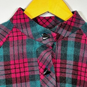 May include: Close-up of a plaid shirt in shades of teal, pink, and black. The shirt features a mandarin collar and three black triangular buttons. The fabric is a woven material, and the shirt is hanging on a wooden hanger.