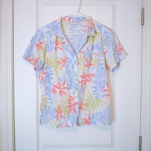 May include: A short-sleeved button-down shirt with a tropical floral pattern. The shirt features a white background with a repeating design of pink, blue, and green leaves. The shirt is hanging on a white hanger against a white door.