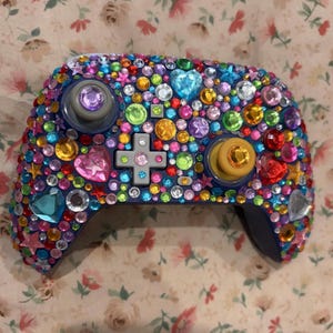 May include: A video game controller covered in a dense arrangement of multicolored, multifaceted rhinestones in various shapes including circles, stars, and hearts. The controller features a gray directional pad and two analog sticks.