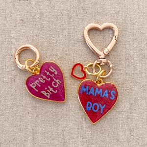May include: Two gold heart-shaped keychains with red enamel. One keychain says "Pretty Bitch" in white lettering and the other says "Mama's Boy" in blue lettering.