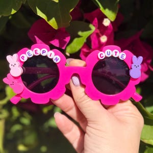 May include: Bright pink sunglasses with a floral design around the lenses. The text "PEEPIN" and "CUTE" are spelled out in white beads. The sunglasses feature pink and purple bunny accents.