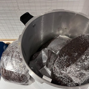May include: A silver pressure cooker containing several large, dark brown, packaged items. The items are wrapped in clear plastic. The cooker is on a white surface, with a blue and silver object visible on the left.