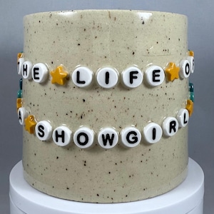 Peut inclure: Un mug cylindrique en céramique avec une finition beige mouchetée. Les mots "THE LIFE OF A SHOWGIRL" sont épelés avec des perles de lettres blanches, entrecoupées de perles en forme d'étoile jaunes. Le mug repose sur une surface blanche.