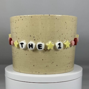 Peut inclure: Un pot en céramique beige moucheté avec une bande de perles qui dit "THE 1". La bande comporte des perles de lettres blanches, des perles étoiles jaunes et des perles roses et rouges. Le pot repose sur une surface blanche.