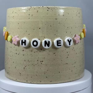 Peut inclure: Un mug en céramique beige moucheté avec une guirlande de perles épelant "HONEY". La guirlande comprend des perles de lettres blanches et des perles colorées en forme de fleurs et de bonbons. Le mug est cylindrique et repose sur une surface blanche.