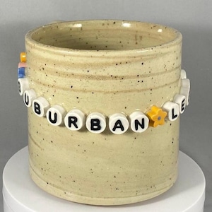 Peut inclure: Un mug en céramique beige moucheté avec l'inscription "SUBURBAN LIFE" en perles de lettres blanches. Le mug est de forme cylindrique et possède une petite anse. Une perle jaune en forme d'étoile est incluse dans le mot "LIFE".