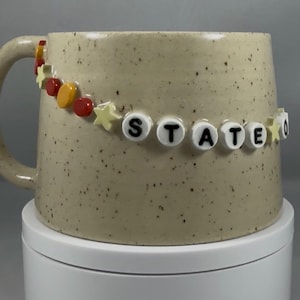 Peut inclure: Une tasse en céramique beige mouchetée avec une grande anse. La tasse est décorée d'une guirlande de perles avec des étoiles, des perles rouges, jaunes et oranges, et le mot "STATE" en perles blanches.
