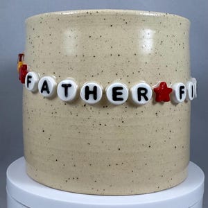 Peut inclure: Un mug en céramique beige moucheté, orné d'une guirlande de perles épelant "FATHER". Les perles sont blanches avec des lettres noires et une étoile rouge. Le mug est posé sur une surface blanche.