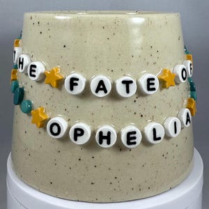 Peut inclure: Un mug en céramique beige avec le texte "THE FATE OF OPHELIA" en perles de lettres blanches. Les perles sont enfilées avec des étoiles jaunes et des perles turquoise. Le mug est sur une surface blanche.