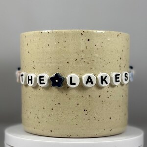 Peut inclure: Un mug en céramique beige moucheté avec une bande de perles qui épelle "THE LAKES". La bande comprend des perles blanches avec des lettres noires et une perle fleur bleu foncé. Le mug est posé sur une surface blanche.
