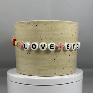 Peut inclure: Un pot de fleurs en céramique beige avec une finition mouchetée. Le pot est orné d'une bande de perles épelant "LOVE STORY" avec des perles de lettres blanches et des accents de fleurs roses. Le pot repose sur un piédestal blanc.