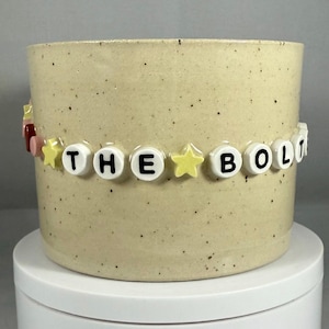 Peut inclure: Un pot de fleurs en céramique beige moucheté avec une bande de perles épelant "THE BOLT". La bande comprend des perles de lettres blanches, des perles étoiles jaunes et des perles d'accent rouges et roses. Le pot est posé sur une surface blanche.
