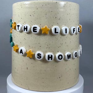 Peut inclure: Un mug cylindrique en céramique avec une finition beige mouchetée. Les mots "THE LIFE" et "A SHOWGIRL" sont épelés avec des perles de lettres blanches, accentuées de perles étoiles jaunes. Le mug repose sur un piédestal blanc.