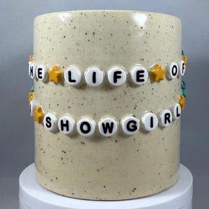 Peut inclure: Un mug en céramique beige moucheté avec l'inscription "THE LIFE OF SHOWGIRL" en perles de lettres blanches. Des perles en forme d'étoiles jaunes et turquoise ornent le texte. Le mug cylindrique est posé sur une surface blanche.