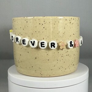Peut inclure: Un pot de fleurs en céramique beige moucheté, orné d'une bande de perles blanches épelant "FOREVER & A". Le pot cylindrique est décoré de petites mouchetures noires. La bande comprend de petites perles en forme de fleurs roses et pêche.