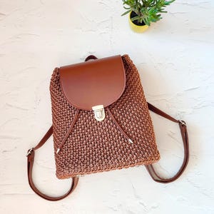 May include: A brown woven backpack with a leather flap and adjustable straps. The backpack has a silver clasp and drawstring closure. A small green plant in a yellow pot is in the background.
