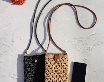 May include: Two rectangular crossbody phone pouches made of crocheted cord, one in black and one in beige. Each pouch features a matching faux leather strap, a snap-button closure with a faux leather tab, and a black smartphone placed beside them.