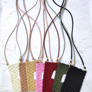Puede incluir: Colección de siete fundas de teléfono de ganchillo en varios colores: crema, beige, dorado, rosa, burdeos, verde oliva y negro. Cada funda tiene una correa a juego y una pequeña etiqueta. Sobre un fondo blanco texturizado.