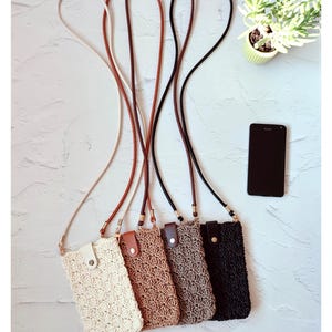 Puede incluir: Cuatro bolsos cruzados para teléfono de ganchillo en crema, beige, marrón y negro, cada uno con una correa a juego. Las bolsas tienen un cierre de botón y se muestran con un teléfono inteligente negro y una pequeña planta en maceta.