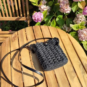 May include: A black crocheted crossbody bag with a rectangular shape and an open-weave texture. The bag features a thin black leather-like strap with an adjustable buckle and a small black flap closure secured with a silver-toned metal snap button.