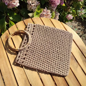 May include: A square-shaped beige crochet handbag featuring a textured open-weave pattern and a circular bamboo handle. The bag is constructed from a light tan cord material and includes a reinforced top border with a consistent stitch design.