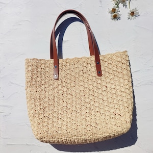 May include: A beige crochet tote bag featuring a repeating shell stitch pattern. The bag has two brown leather straps attached with silver-toned metal rivets. The texture of the woven material is visible throughout the body of the bag.