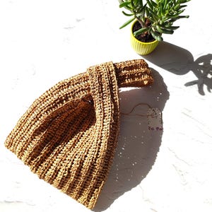May include: A tan, woven straw-like handbag featuring a unique knot-style handle design. The bag has a textured, ribbed appearance and is accompanied by a small, rectangular paper tag attached with a thin string, resting near a small green succulent.