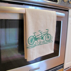 May include: A tan kitchen towel with a green illustration of a tandem bicycle with a floral design. The towel is hanging on a silver oven door.
