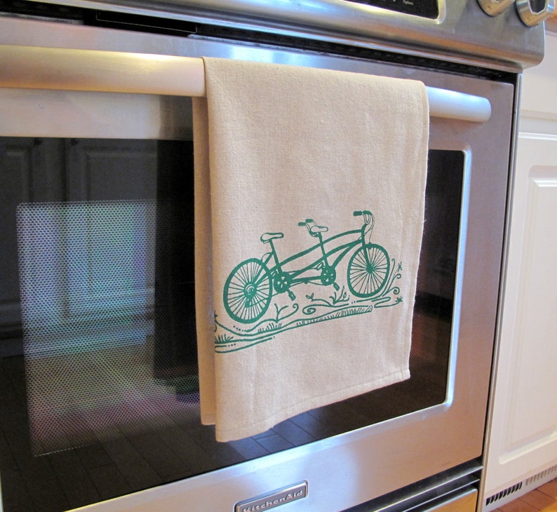 May include: A tan kitchen towel with a green illustration of a tandem bicycle with a floral design. The towel is hanging on a silver oven door.