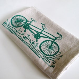 May include: A green tandem bicycle printed on a beige linen tea towel. The bicycle has two seats and handlebars.
