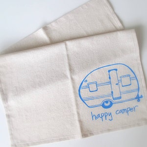 Puede incluir: Un pa&ntilde;o de cocina de algod&oacute;n blanco con una ilustraci&oacute;n azul de una caravana vintage y el texto "happy camper".
