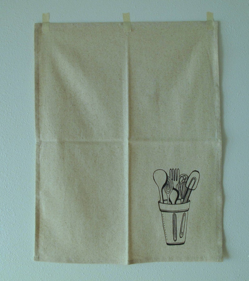 Pu&ograve; includere: Un asciugamano da cucina beige con un'illustrazione in bianco e nero di utensili da cucina in una tazza.