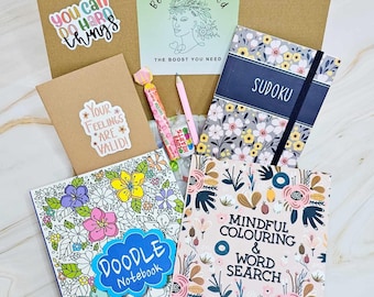 Può includere: Una confezione regalo contenente vari articoli. Include un "Doodle Notebook", un libro "Mindful Colouring & Word Search", un quaderno "Sudoku", adesivi, una matita e un bastoncino di caramelle. La scatola è marrone e gli articoli hanno motivi floreali.