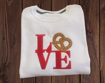 Op de afbeelding: Wit sweatshirt met het woord "LOVE" in rode letters en een pretzelontwerp in plaats van de "O". De trui heeft een ronde hals en een groene kraagafwerking. Het pretzelontwerp is bruin. Het kledingstuk ligt op een houten oppervlak.