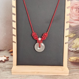 Könnte beinhalten: Eine rote Schnurkette mit einem weißen Jade-Anhänger und roten Perlen. Die Kette wird auf einem Holzständer präsentiert.