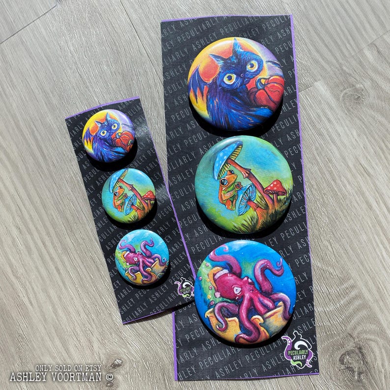 Op de afbeelding: Set van vijf 3,8 cm grote knoopsluitingen met handgeschilderde illustraties. De knoopsluitingen tonen een zwarte vleermuis met rode ogen, een kikker die op een paddenstoel zit, een paarse octopus in een pot, een zwarte kat met rode ogen en een kikker die op een paddenstoel zit. De knoopsluitingen bevinden zich op een zwarte achtergrond met witte tekst die "Peculiarly Ashley" luidt.