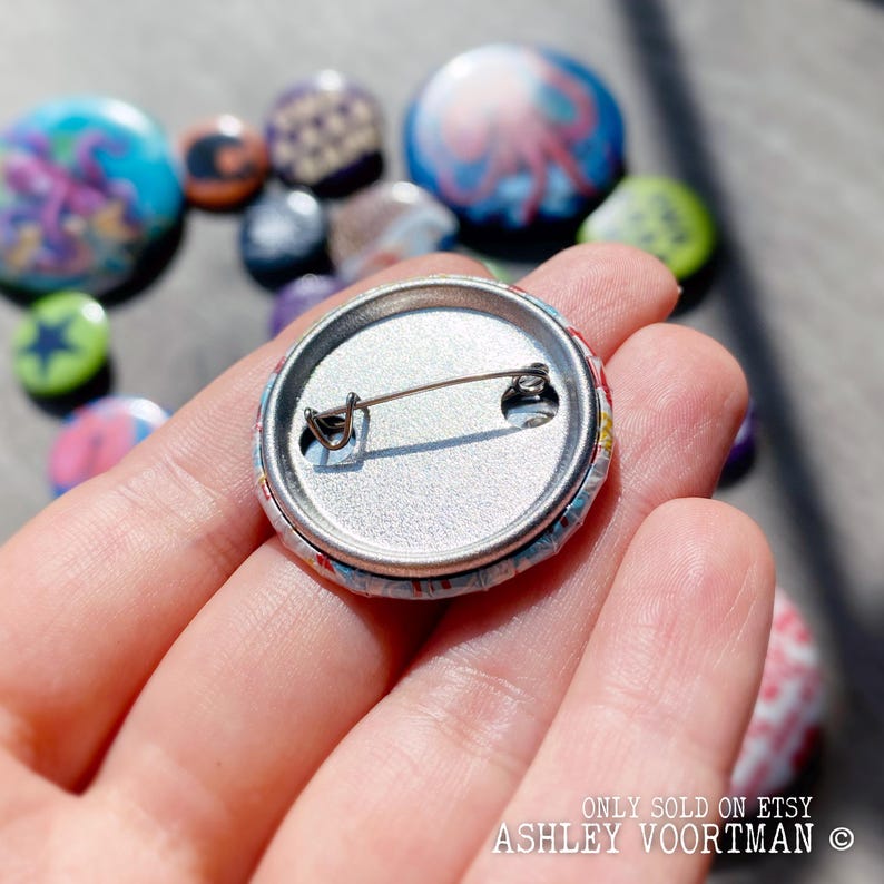 Op de afbeelding: De achterkant van een kleurrijke knoop met een pin aan de achterkant. De knoop wordt vastgehouden door een hand. De tekst op de knoop luidt "ONLY SOLD ON ETSY ASHLEY VOORMAN &copy;".
