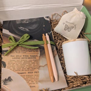 Pode incluir: Uma caixa de presente com um caderno, canetas, uma vela e uma pequena bolsa. O caderno tem uma imagem de lente de câmera e o texto "FOCUS FREE LENS MADE IN JAPAN". As canetas são bege e rosa. A bolsa tem um logotipo de borboleta.