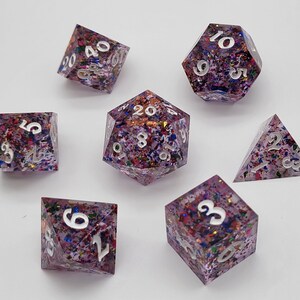 May include: A set of seven polyhedral dice made of clear resin filled with multicolored glitter and foil flakes. Each die features sharp edges and engraved numbers painted in white, including a d20, d12, d10, d8, d6, d4, and a percentile die.