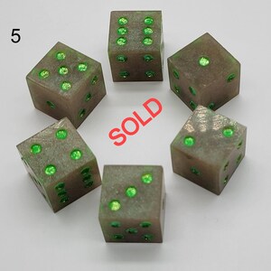 May include: A set of six translucent gray dice with bright green glitter pips. The number 5 is printed in the upper left corner, and the word SOLD is printed in red in the center of the arrangement of the dice on a plain white background.