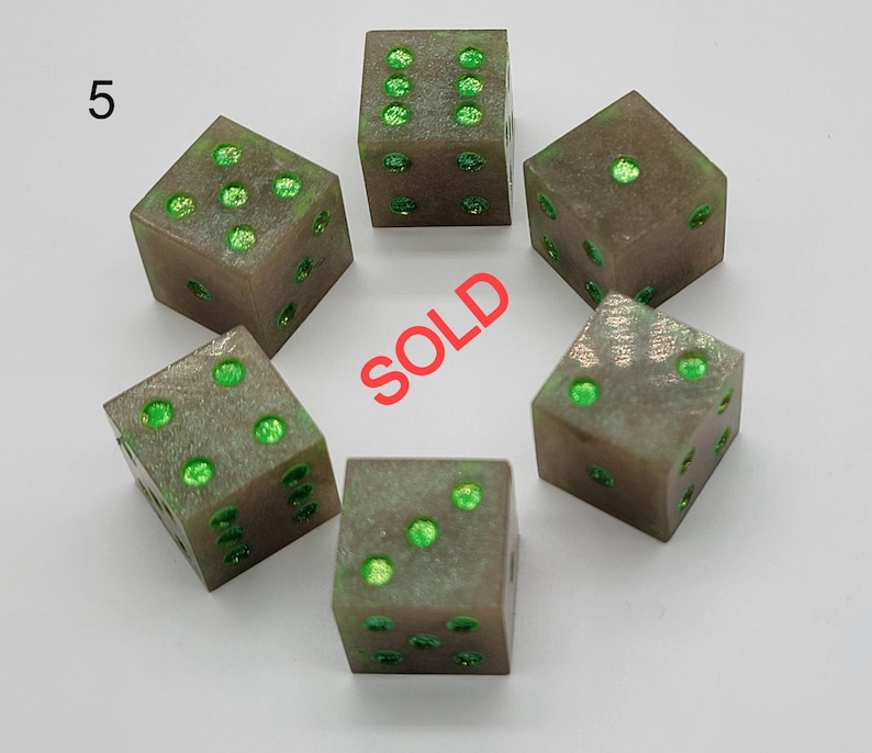 May include: A set of six translucent gray dice with bright green glitter pips. The number 5 is printed in the upper left corner, and the word SOLD is printed in red in the center of the arrangement of the dice on a plain white background.