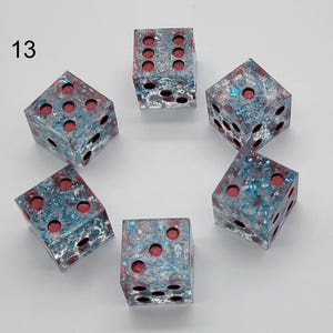 May include: A set of six translucent dice labeled with the number 13, featuring a clear resin body embedded with blue and silver metallic flakes and deep red circular pips on each face, arranged in a circular pattern on a plain white background.
