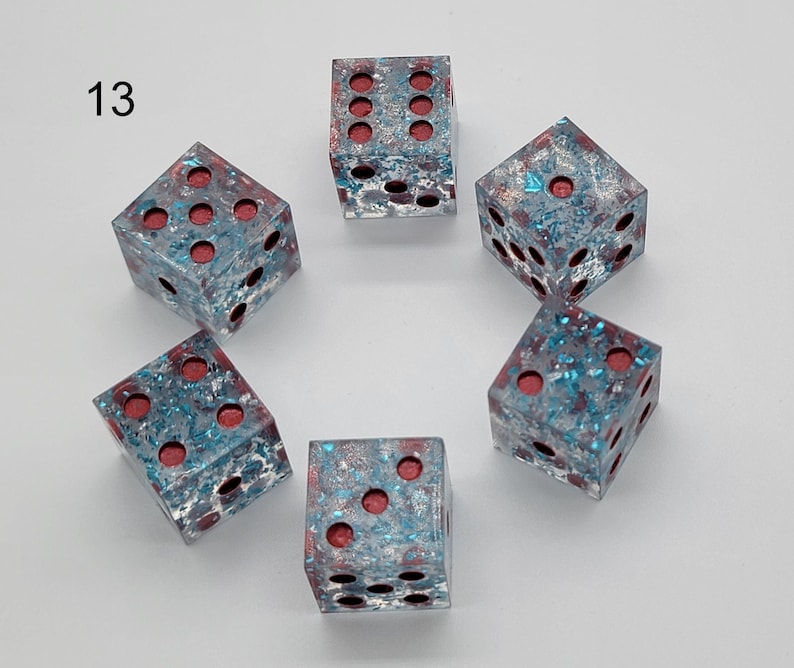 May include: A set of six translucent dice labeled with the number 13, featuring a clear resin body embedded with blue and silver metallic flakes and deep red circular pips on each face, arranged in a circular pattern on a plain white background.