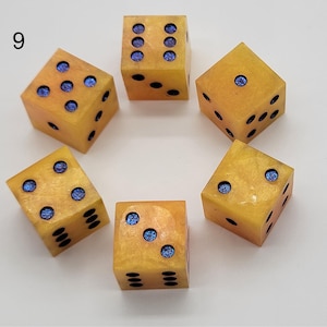 May include: A set of six yellow dice arranged in a circle, each featuring blue glitter-filled pips. The dice have a smooth, semi-translucent finish with a marbled appearance. The number nine is printed in black in the upper left corner of the image.