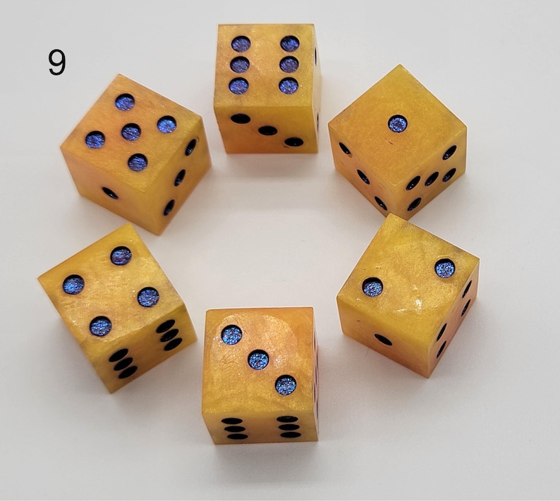 May include: A set of six yellow dice arranged in a circle, each featuring blue glitter-filled pips. The dice have a smooth, semi-translucent finish with a marbled appearance. The number nine is printed in black in the upper left corner of the image.